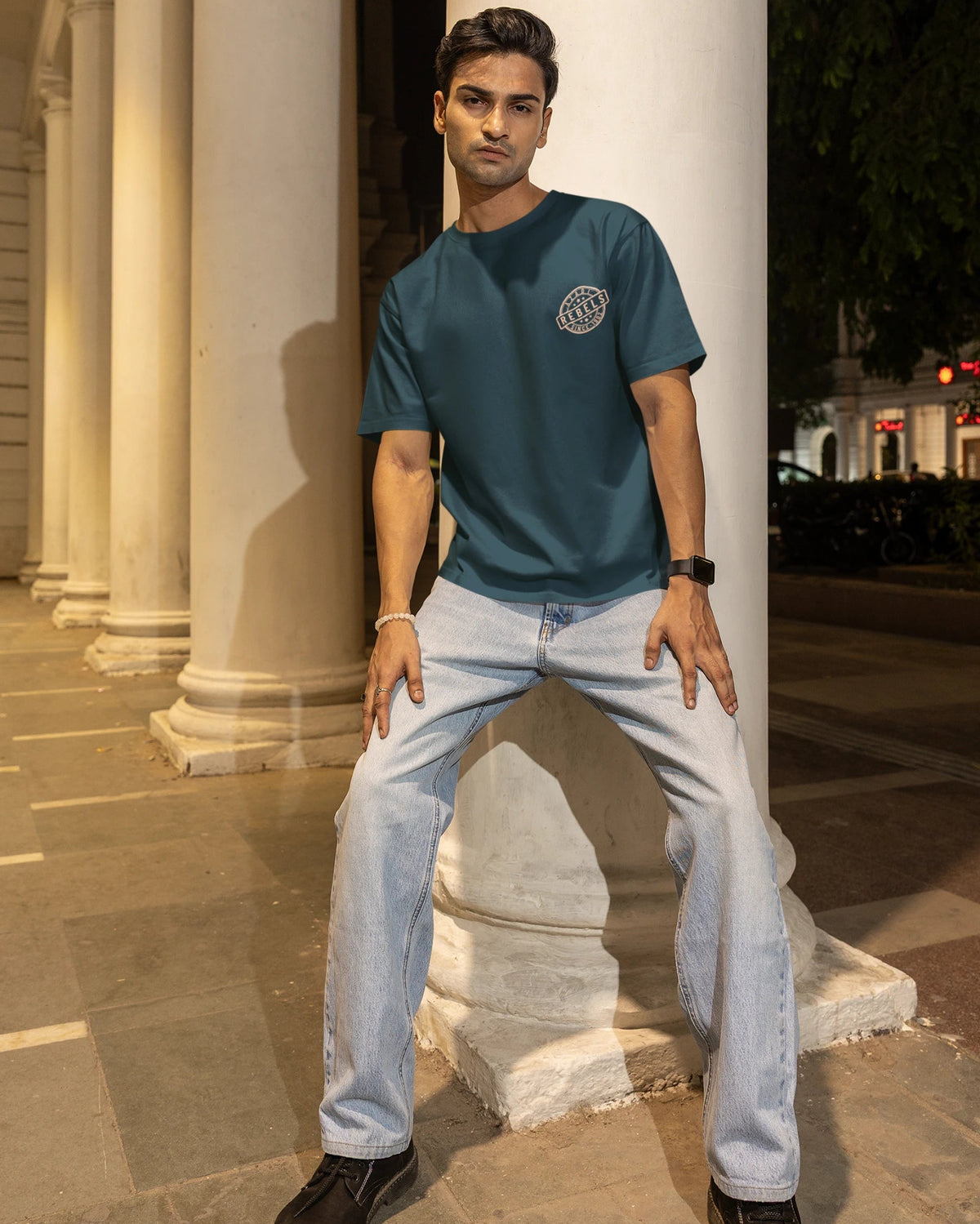 A man posing in a teal blue t-shirt with a circular graphic on the left chest, paired with light wash jeans and black shoes, standing in front of a large white column at night.