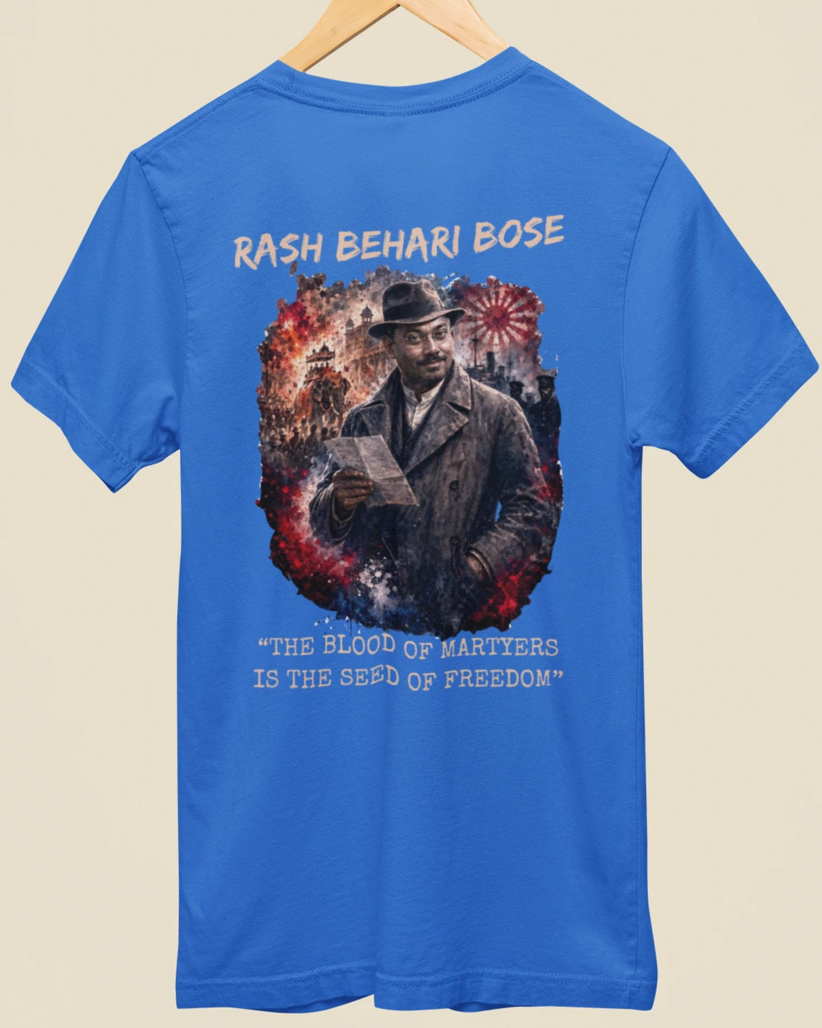 A blue short-sleeve t-shirt hanging on a wooden hanger against a light beige background. The back of the shirt features a large graphic print of a man in a hat and coat, surrounded by red and white fireworks, with the text 'RASH BEHARI BOSE' above and 'THE BLOOD OF MARTYRS IS THE SEED OF FREEDOM' below.