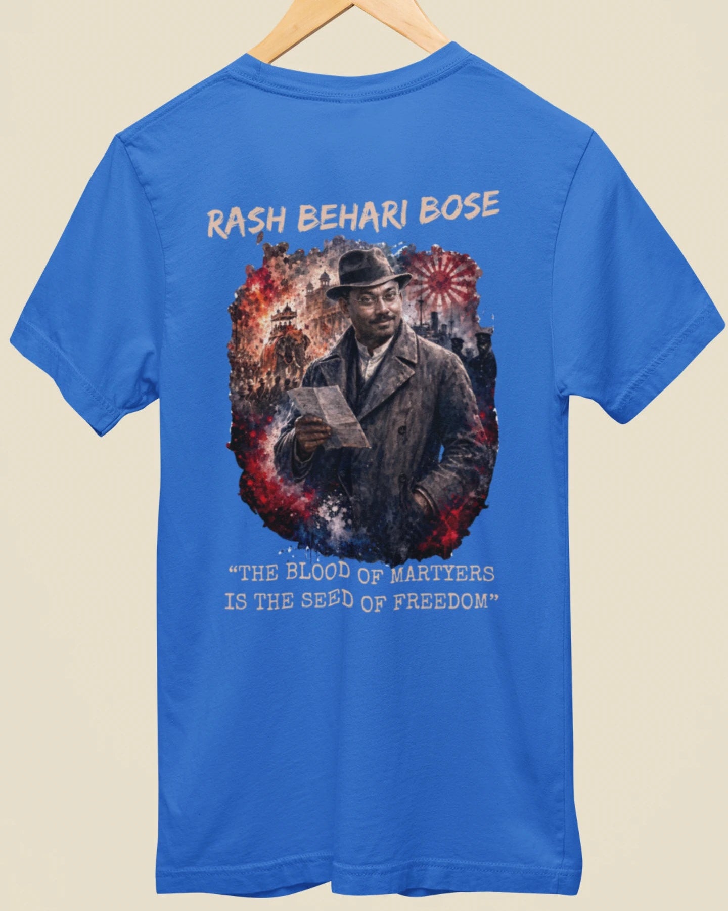 A blue short-sleeve t-shirt hanging on a wooden hanger against a light beige background. The back of the shirt features a large graphic print of a man in a hat and coat, surrounded by red and white fireworks, with the text 'RASH BEHARI BOSE' above and 'THE BLOOD OF MARTYRS IS THE SEED OF FREEDOM' below.