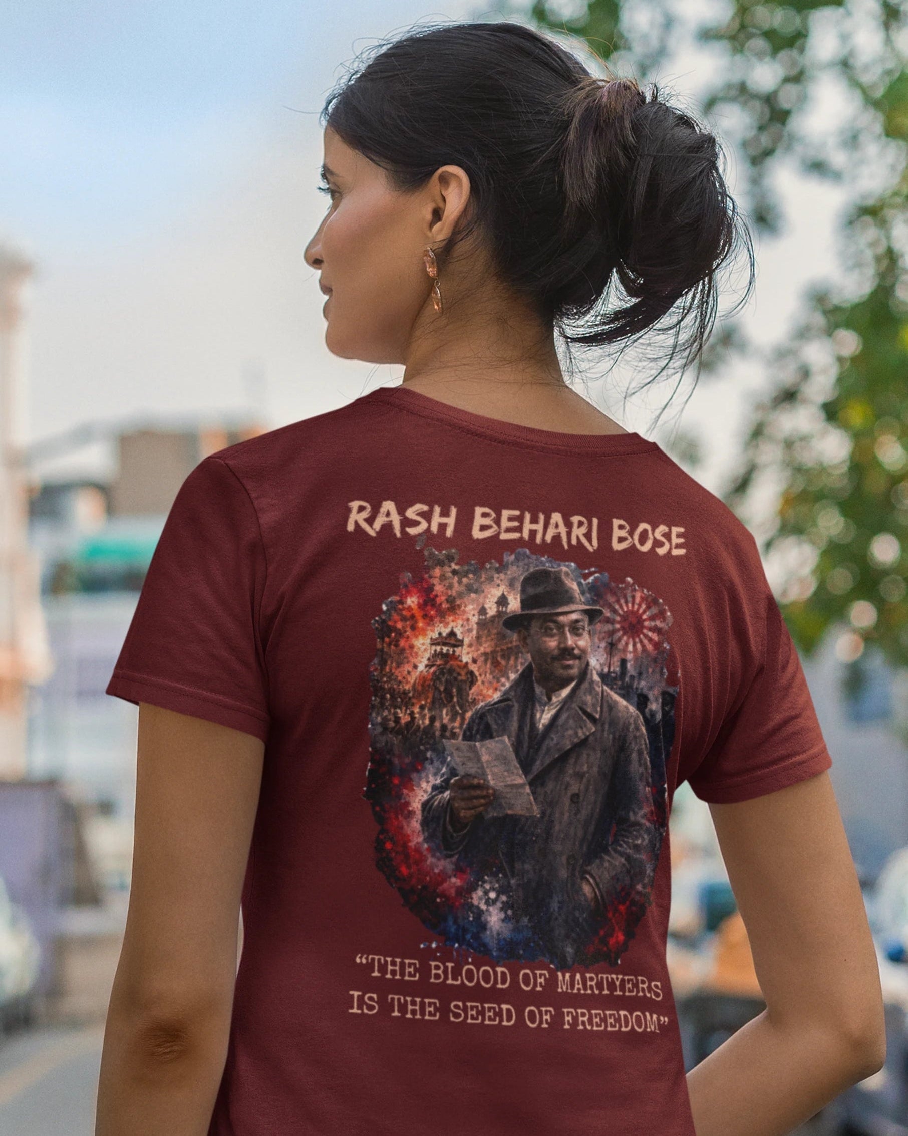 A woman shown from behind wearing a maroon short-sleeve t-shirt with a large graphic print on the back. The graphic features the text 'RASH BEHARI BOSE' and 'THE BLOOD OF MASTERS IS THE SEED OF FREEDOM' surrounding an illustration of a man in a hat.
