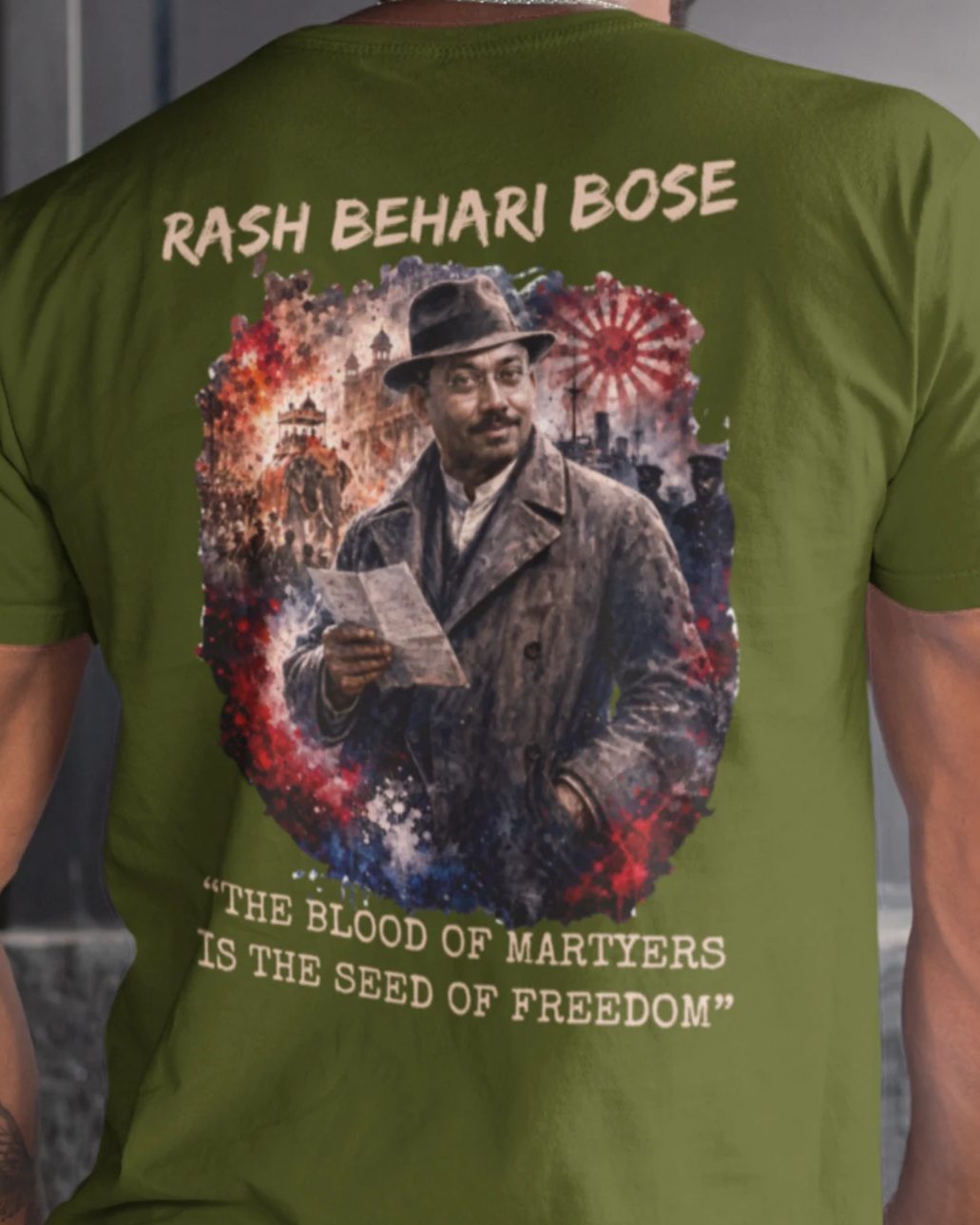 An olive green short-sleeve t-shirt shown from the back on a model, featuring a large graphic print of Rash Behari Bose with the text 'RASH BEHARI BOSE' and 'THE BLOOD OF MARTYRS IS THE SEED OF FREEDOM'.