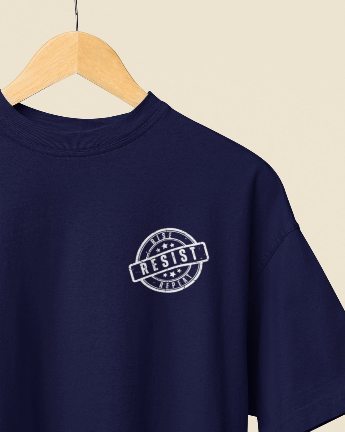 A navy blue short-sleeve t-shirt hanging on a wooden hanger against a light beige background. The shirt features a circular white graphic on the left chest with the word 'RESIST' inside a stamp-like border.
