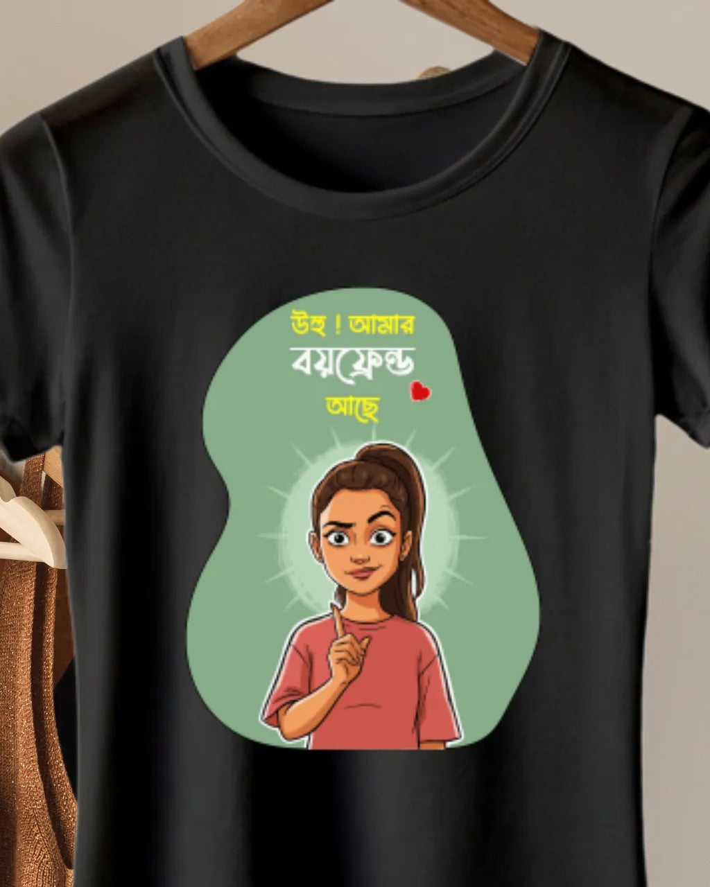 A black short-sleeve t-shirt hanging on a wooden hanger, featuring a cartoon-style graphic of a woman with Bengali text inside a light green pear-shaped outline. Other hangers and garments are visible in the background.