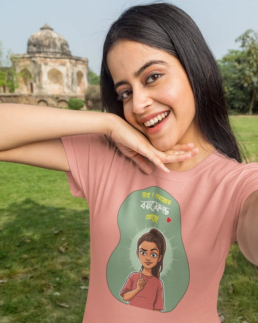 A woman taking a selfie outdoors wearing a light pink t-shirt with a green graphic print of a cartoon character and Hindi text. A grassy park and a historic stone structure are visible in the background.