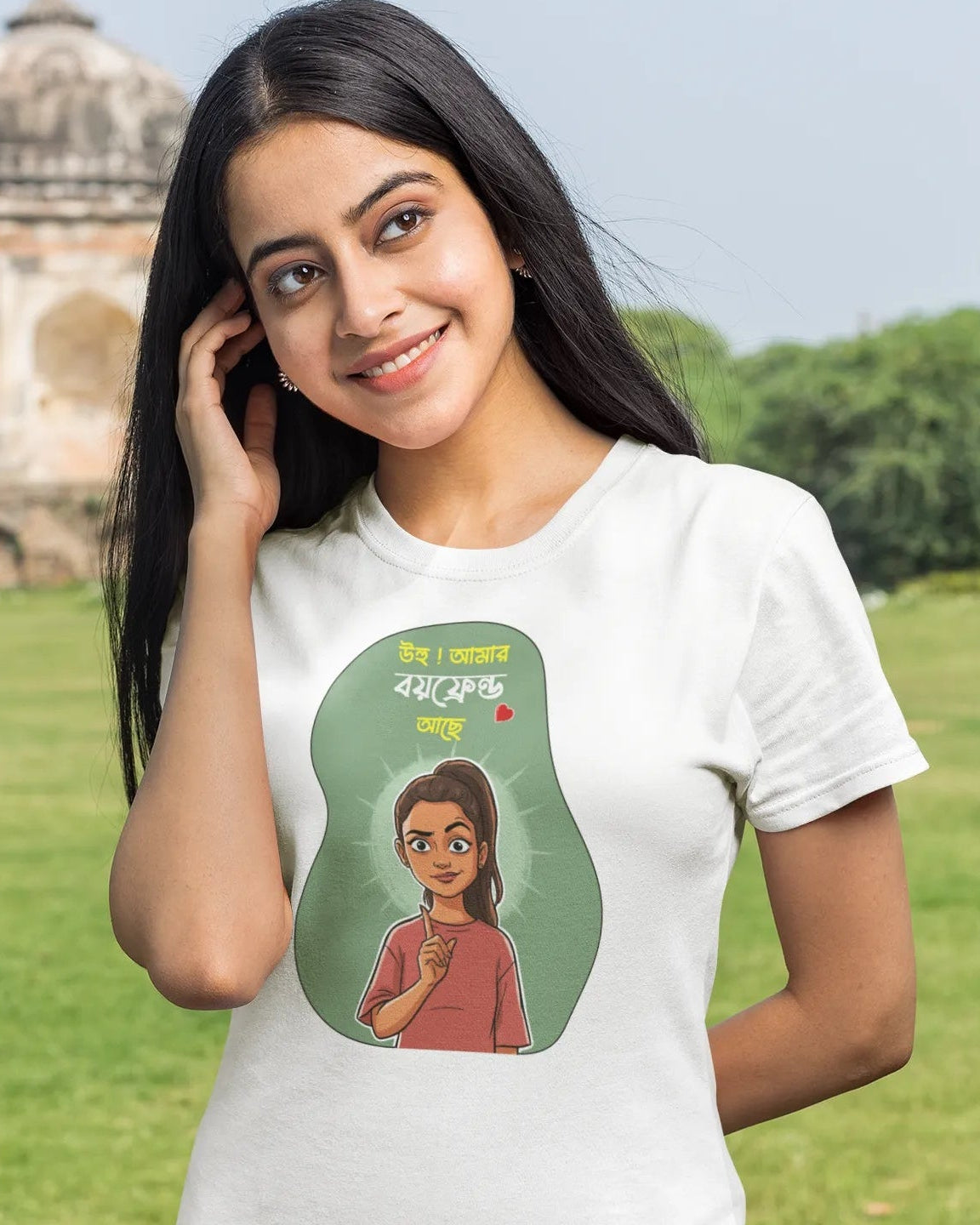 A woman wearing a white graphic t-shirt with a cartoon illustration of a girl and Bengali text, standing in a grassy park with a domed stone structure in the background.
