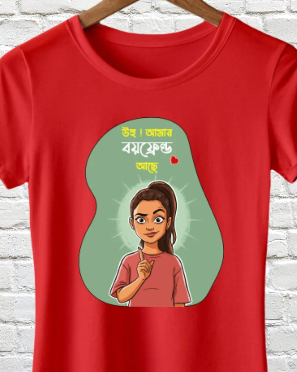 A red short-sleeve t-shirt hanging on a wooden hanger against a white brick wall. The shirt features a graphic print with a green pear-shaped outline containing a cartoon illustration of a woman and Bengali text.