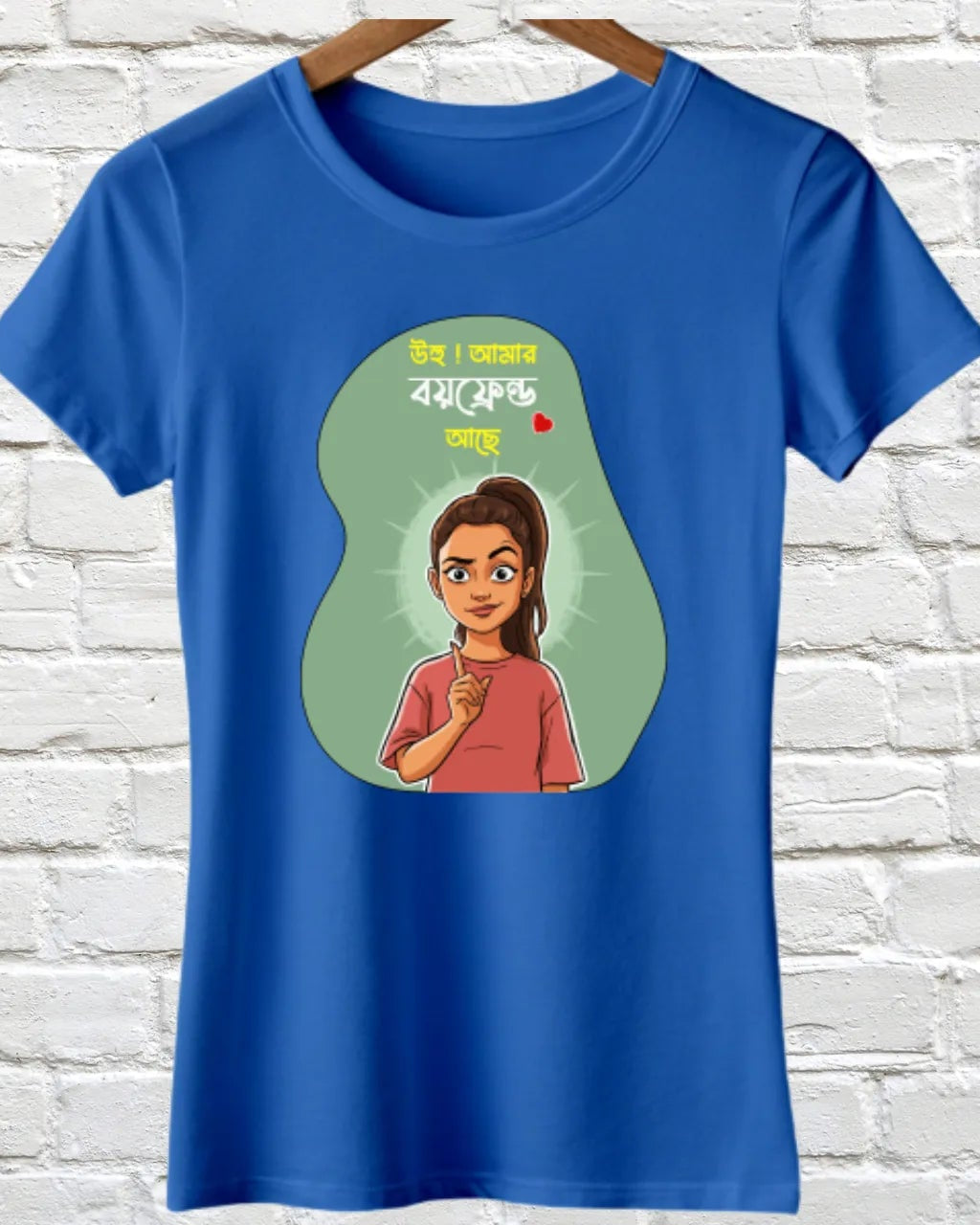 A royal blue short-sleeve t-shirt hanging on a wooden hanger against a white painted brick wall. The front of the t-shirt features a graphic print with a green teardrop shape containing an illustration of a woman and stylized text.