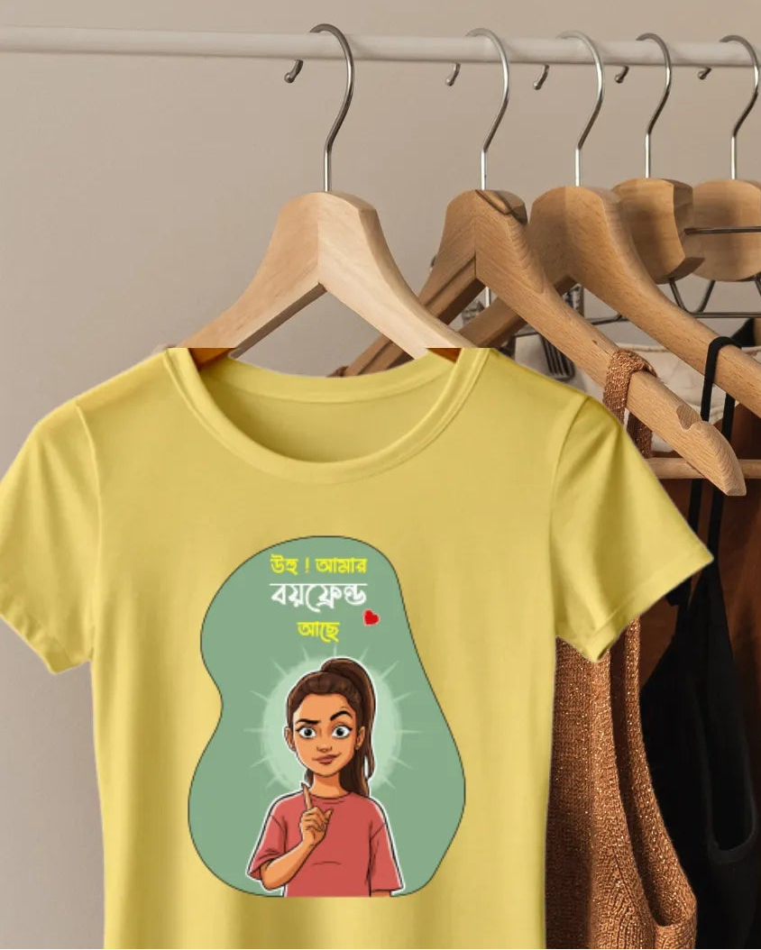 A yellow graphic t-shirt hanging on a wooden hanger on a clothing rack, with other clothes and a straw hat in the background against a plain beige wall.
