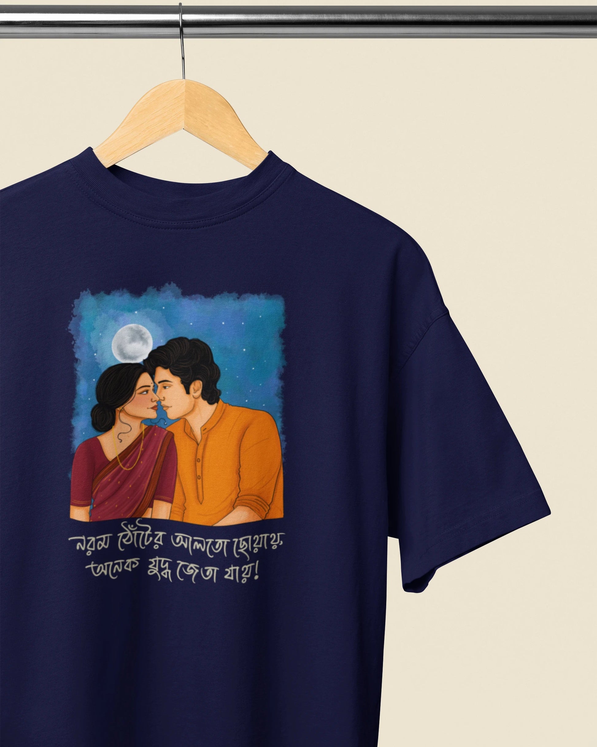A navy blue short-sleeve t-shirt hanging on a wooden hanger against a light beige background. The shirt features a colorful graphic print of a couple kissing under a full moon, with Bengali text written below.
