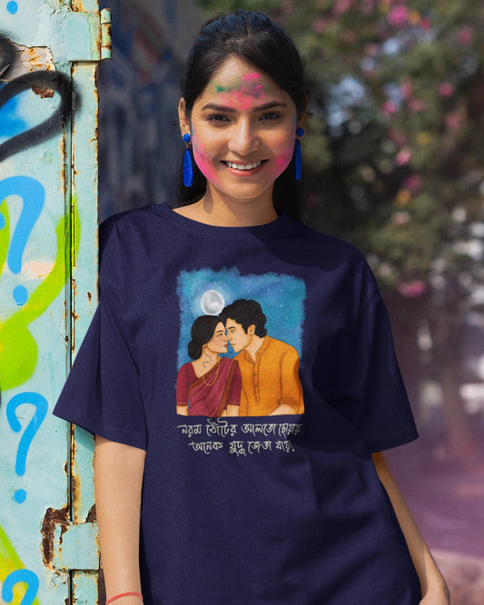 A woman wearing a navy blue graphic t-shirt with a colorful illustration of a couple and Bengali text, standing next to a weathered blue-green door.